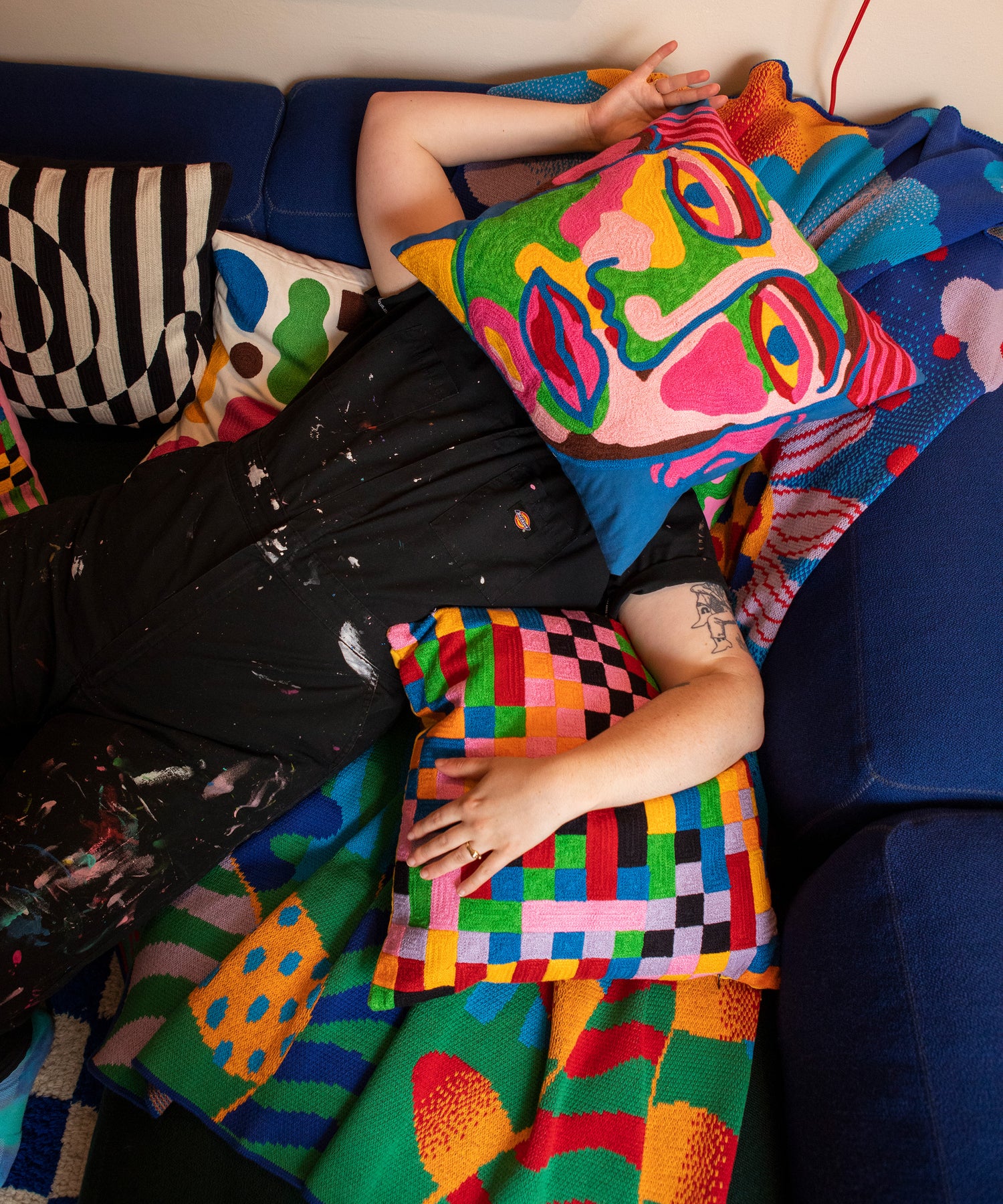 Detail of a person laying on a couch on top of the Best Buds Blanket with the Portrait Pillow covering their face and the Pixels Pillow resting under their left arm.