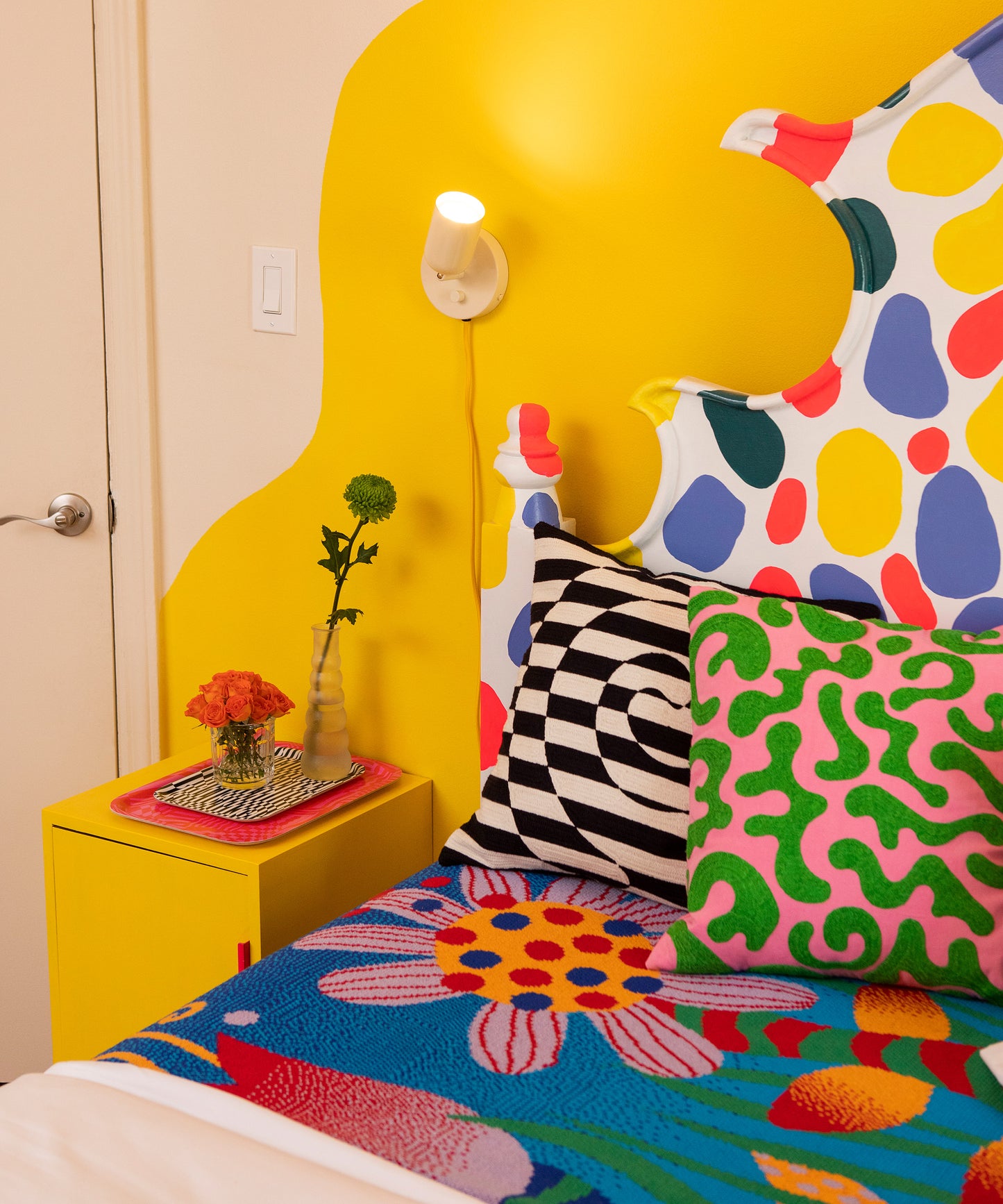 Detail of a bed with the Best Buds Blanket and the Dazzle Pillow sitting on top of it. Sitting on the right of the Dazzle Pillow is the Silly Squiggle Pillow and to the left is a yellow nightstand with the Wiggles and Waves and Dazzle Trays stacked.